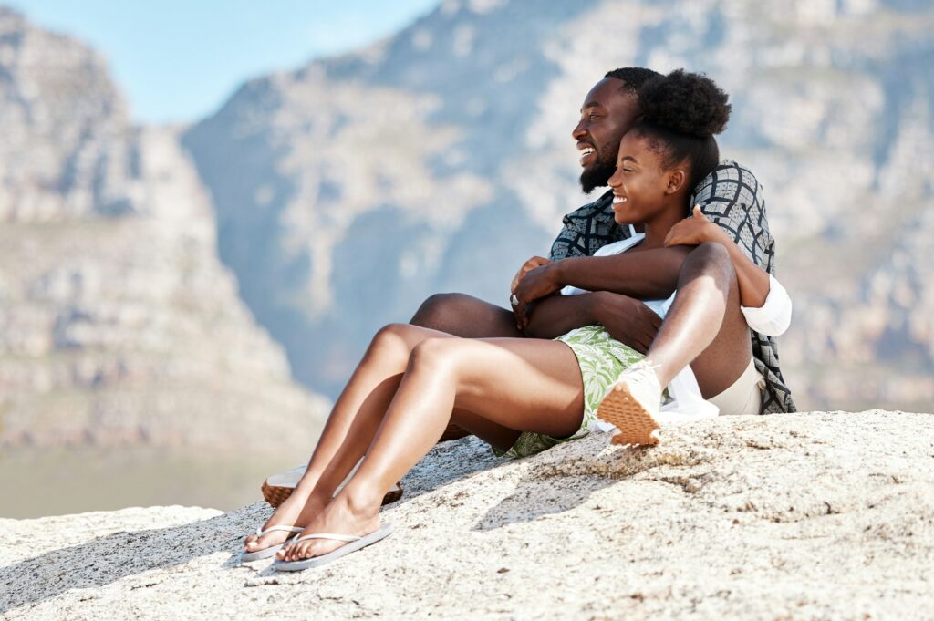Beach, black couple and happy woman and man on rock with South Africa mountain background on summer